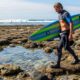 Surfer wearing protective reef boots walking across sharp coral and rocky beach terrain